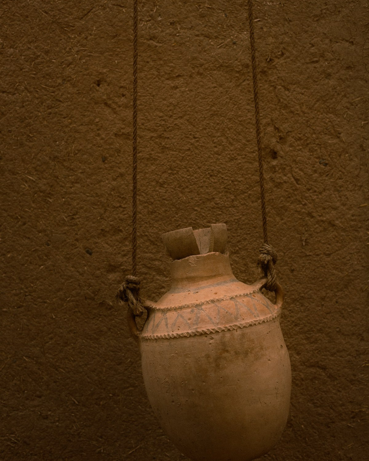 A terracotta water vessel hung on ropes against a clay wall