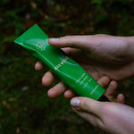 Two hands holding the Rain Wood Hand Cream tube on a forest floor